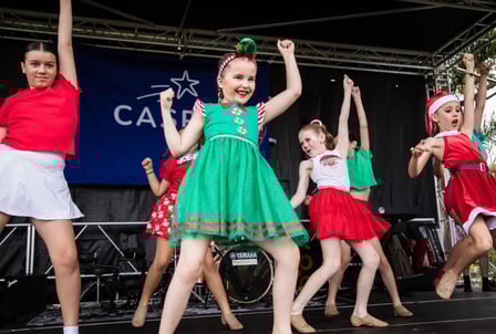 A group of girl's dressed in festive green and red costumes dance energetically on a stage in front of a blue banner with a shooting star that reads CASPA 