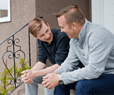 A white teenage boy with light hair sits on the front steps of a house with a white adult male. They are engaged in comfortable conversation. 