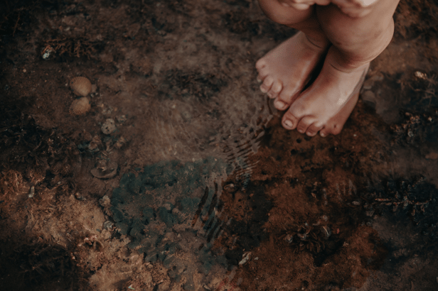 The bare feet of a young Aboriginal person are the only part of them visible. They stand in a rocky creek with clear water.