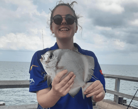 A smiling young person in blue stands on a jetty holding a fish they caught.