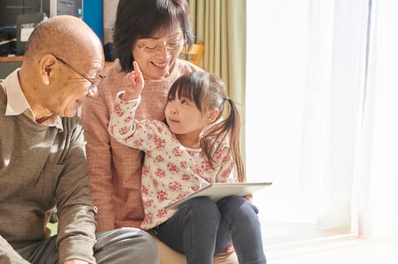 An older Asian man and woman wearing glasses and smiling, sit engaging with a young Asian girl with pigtails using a tablet.